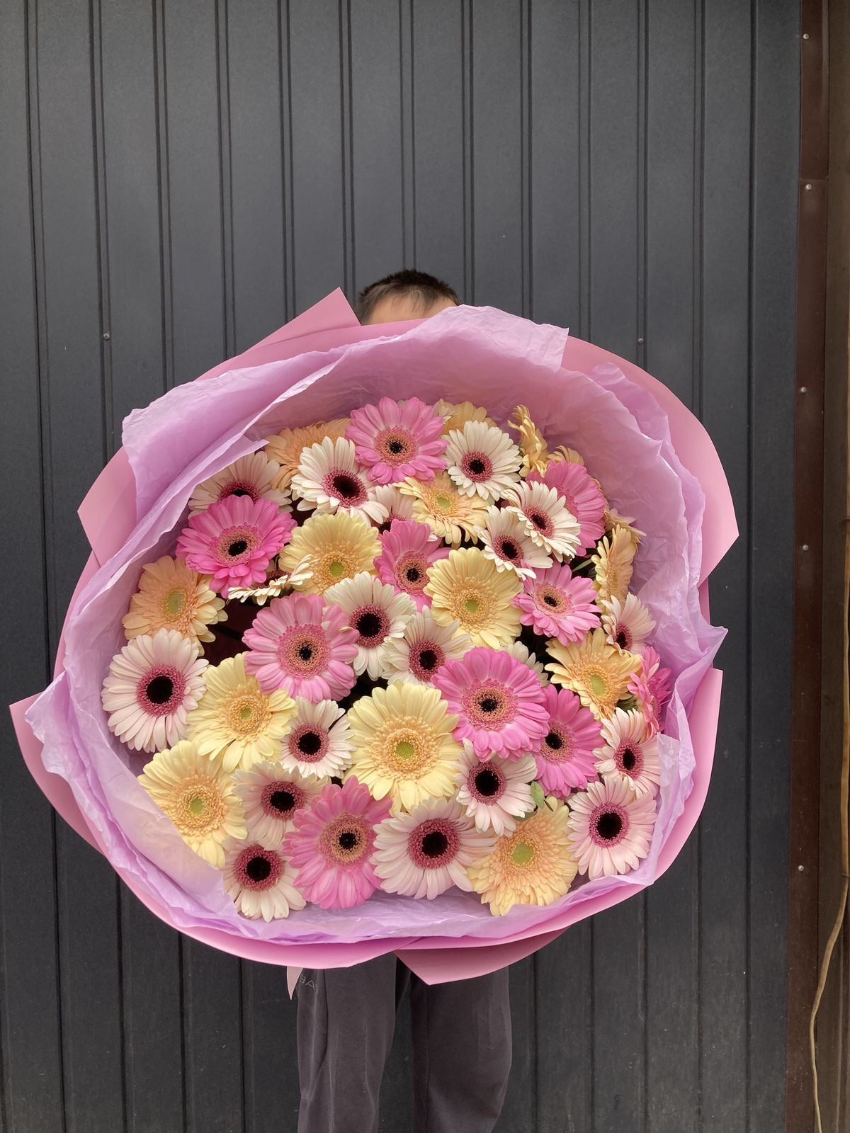 Bouquet Cosette de gerberas roses et jaunes porté devant un mur gris, photo réelle