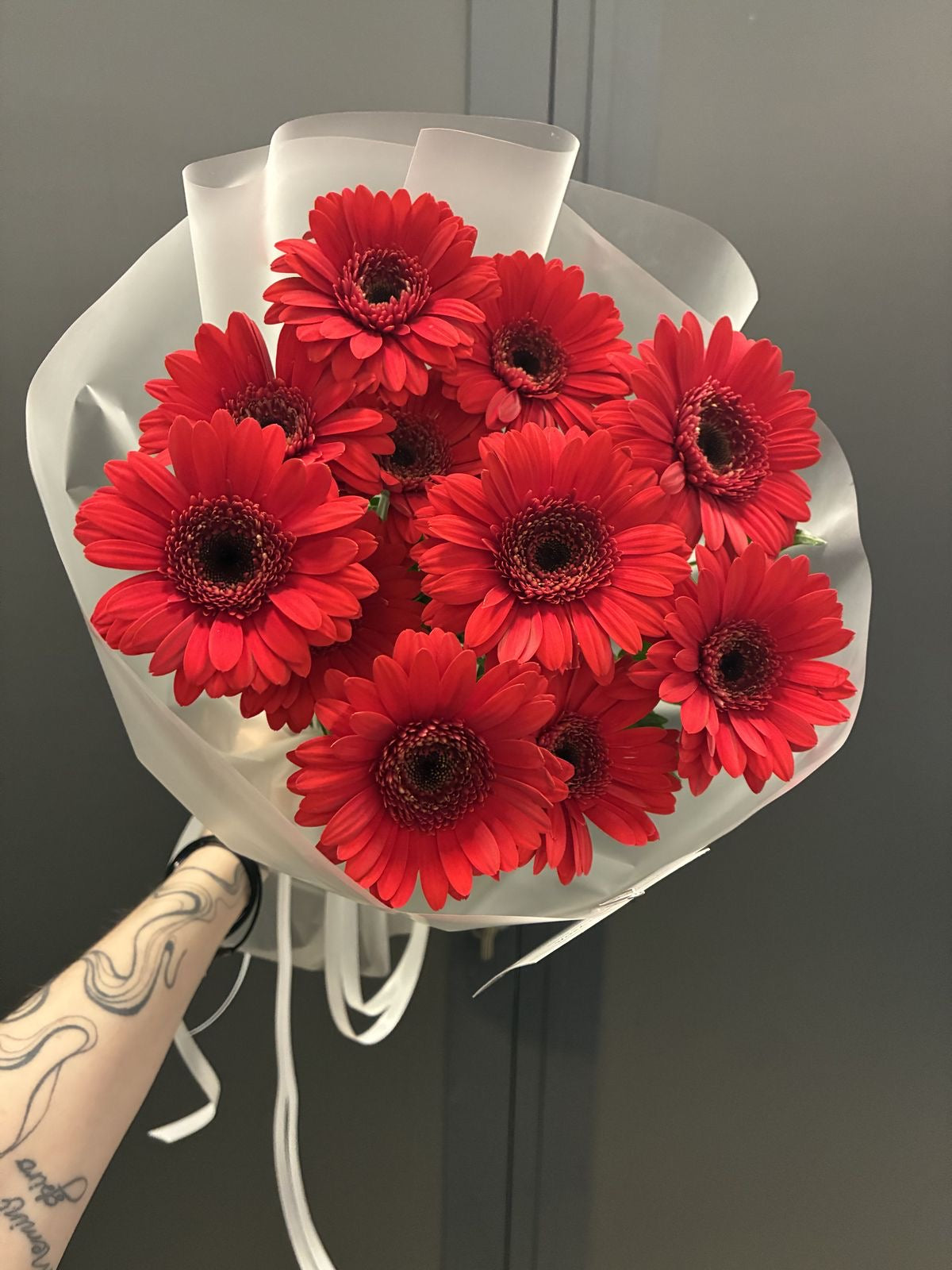 Bouquet Iman de gerberas rouges tenu à la main devant un mur gris, photo réelle