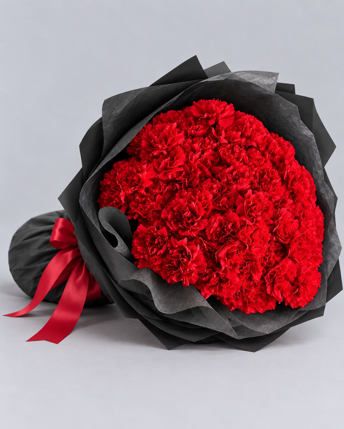Bouquet Mimi d'œillets rouges posé de côté, papier noir et ruban rouge