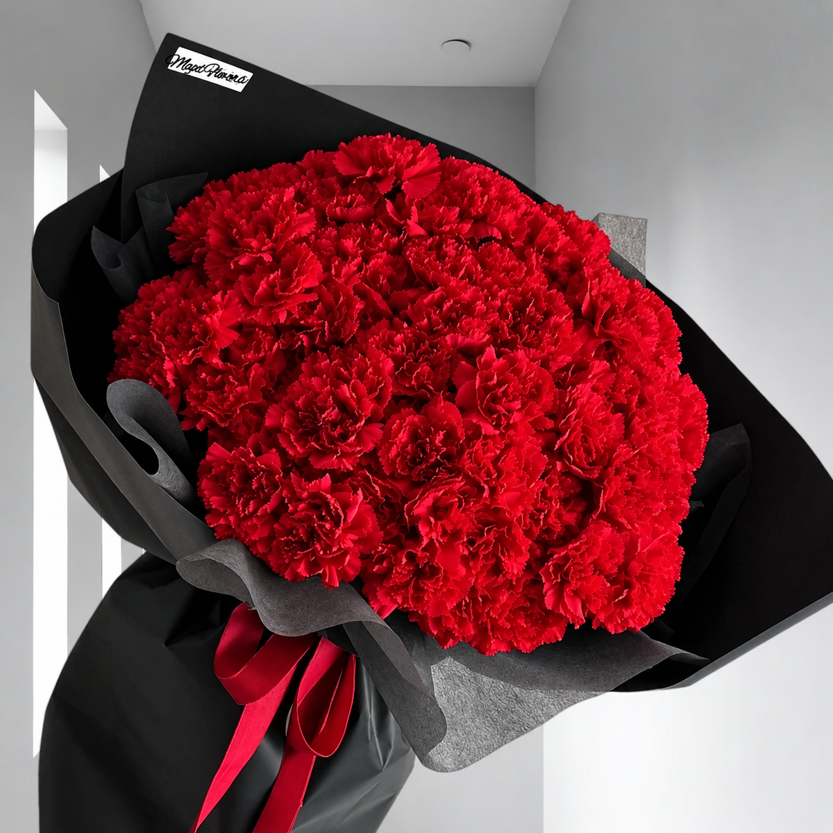 Bouquet Mimi d'œillets rouges avec papier noir et ruban rouge devant une fenêtre