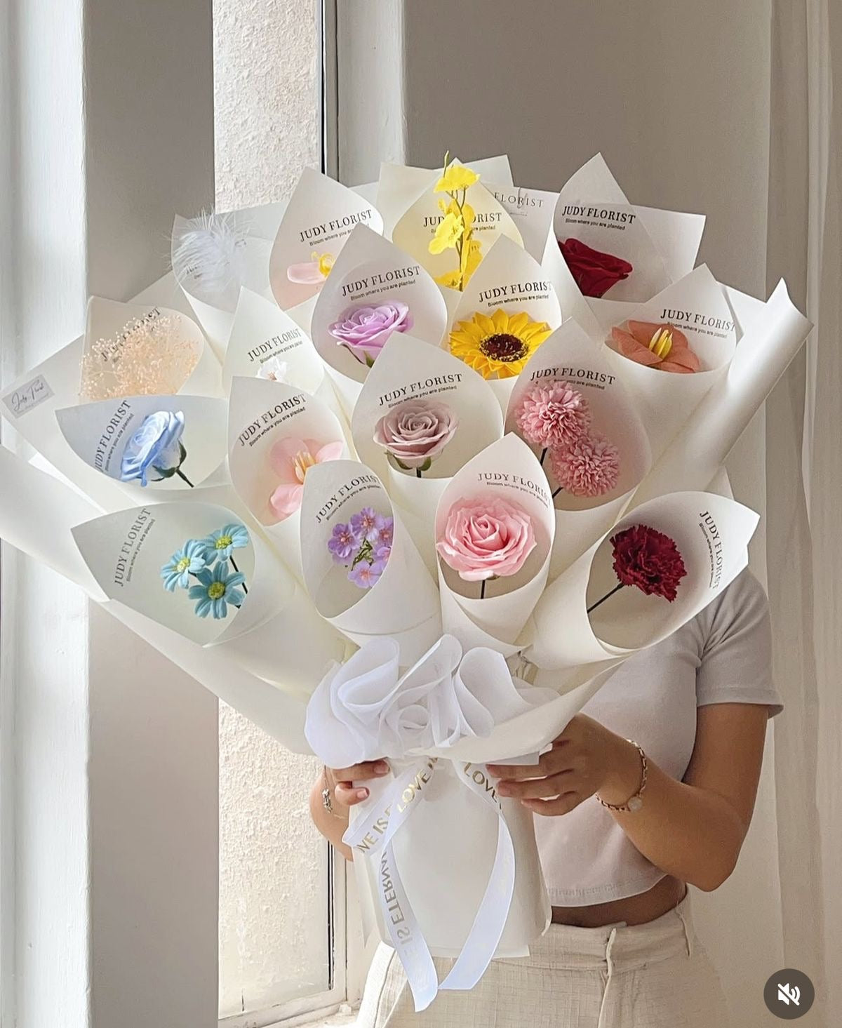 Bouquet CAPRICE de fleurs fraîches multicolores, fleurs enveloppées une à une dans du papier blanc, tenu à deux mains