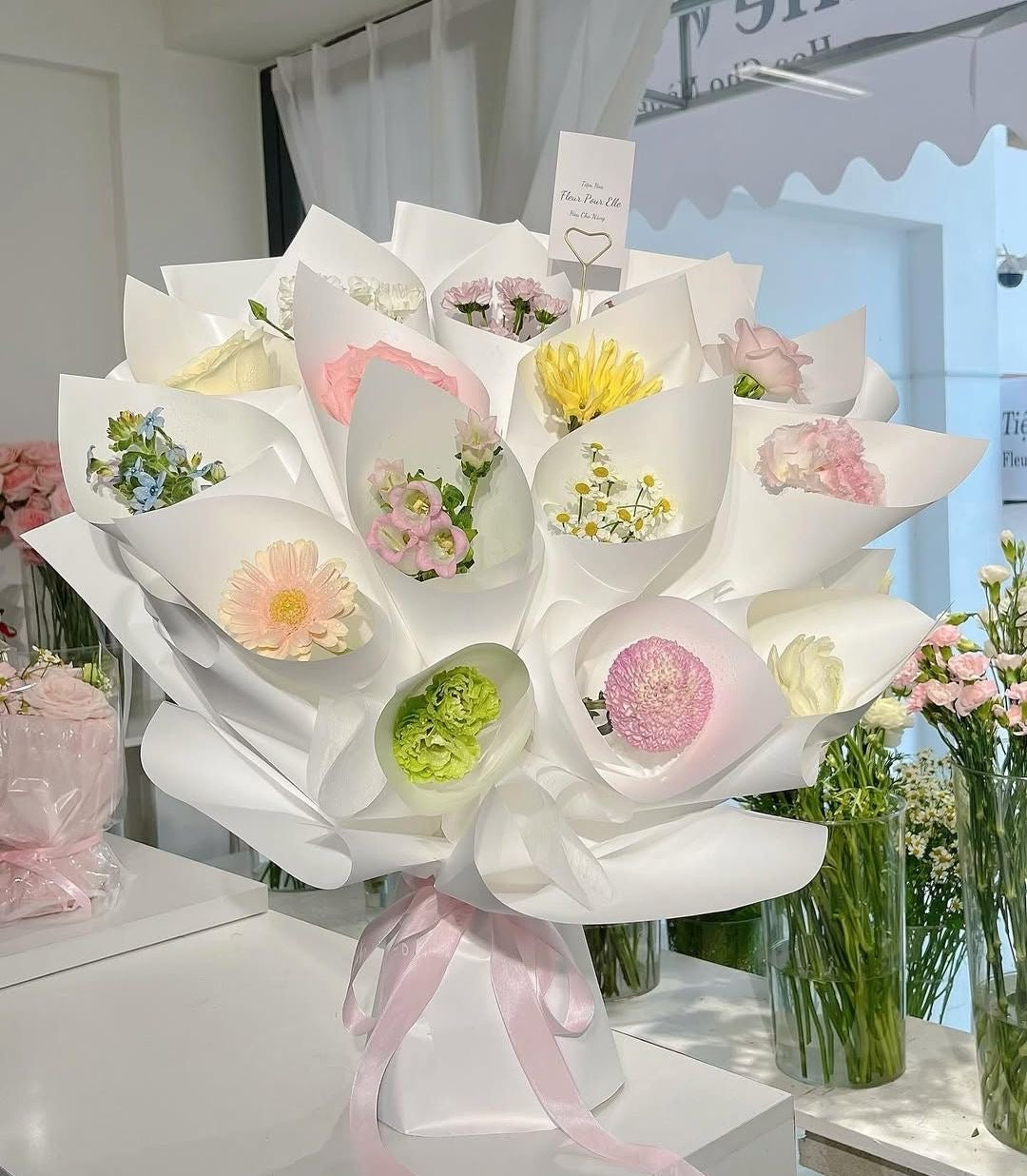 Bouquet CAPRICE de fleurs fraîches multicolores en emballage blanc avec ruban rose, posé sur un comptoir
