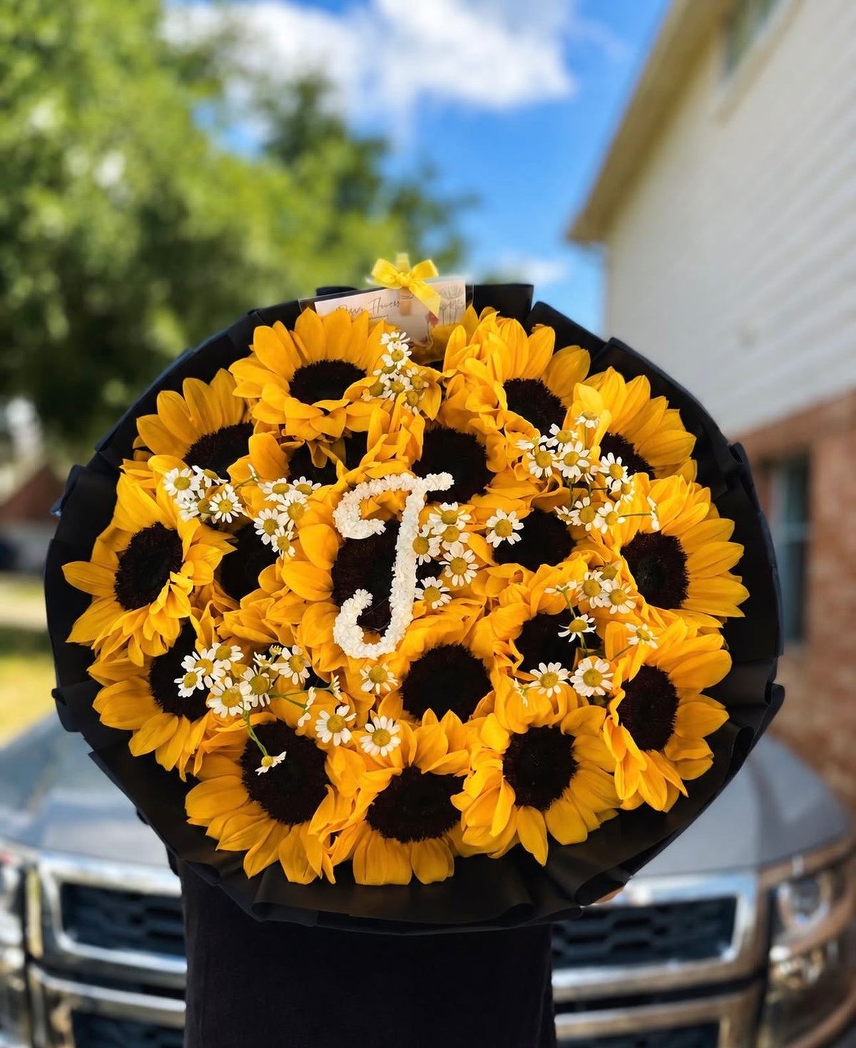 Bouquet Chloé de tournesols avec lettre J tenu en extérieur, papier noir