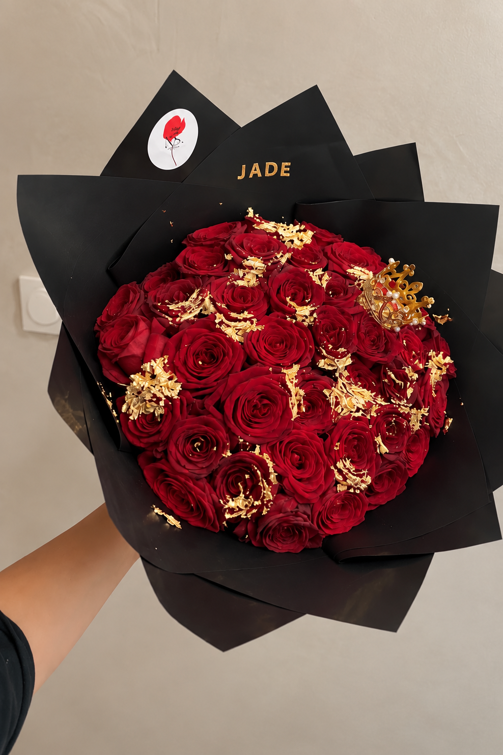 Bouquet ORNELA de roses rouges fraîches et feuilles d'or avec emballage noir, tenu à la main
